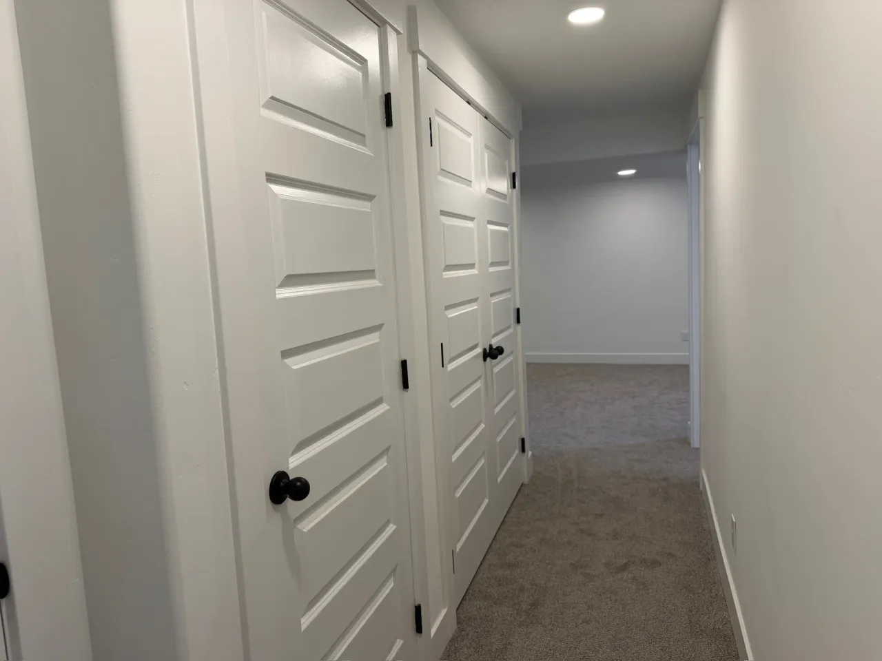 Finished basement hallway with white shaker doors, black hardware, and carpet
